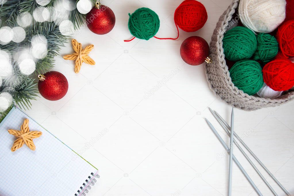 Green and red yarn in a knitted gray basket, a notebook for notes, knitting needles and a spruce branch on a white background. Home comfort and christmas concept. Women's and men's hobby knitting.