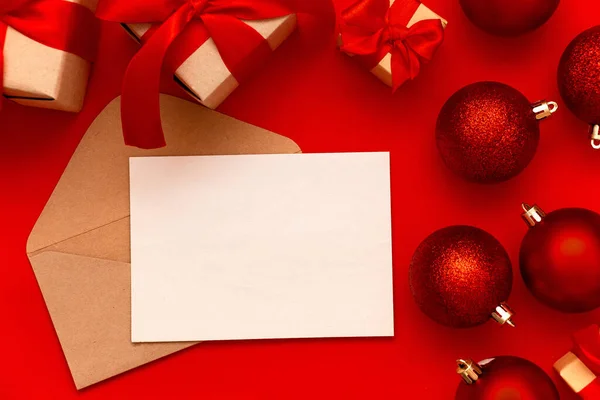 Blank paper sheet, mail envelope and red Christmas balls on a red background with copy space. New Year composition. Flat lay, top view. Minimalistic style.