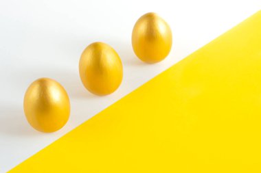 Beyaz-sarı arka planda üç altın rengi yumurta. Minimalist kompozisyon. Paskalya bayramı kartı.