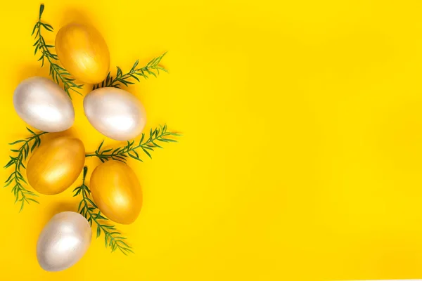 Eggs of silver and gold, green twigs from the left edge of the image on a plain yellow background. Easter holiday card. Copyspace.