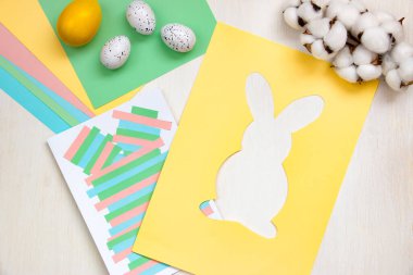 Multi-colored sheets, scissors, glue, Easter eggs, cut out rabbit pattern on a yellow sheet of paper on a light table. DIY concept. Step by step instructions, step 4.
