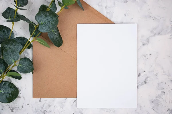 Blank white card, envelope and eucalyptus branch on white marble background. Blank invitation. Flat lay.