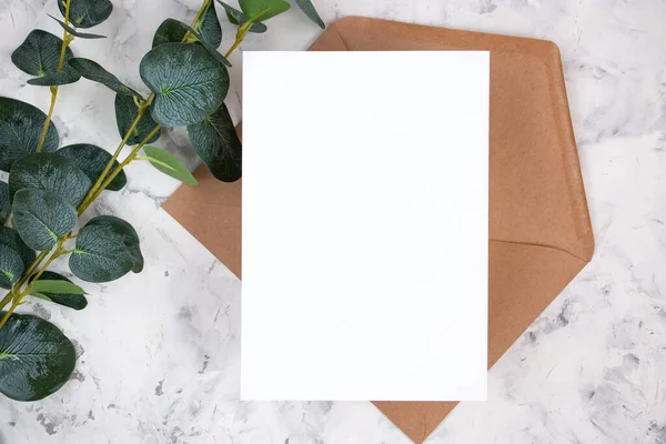 Blank white card, envelope and eucalyptus branch on white marble background. Blank invitation. Flat lay.