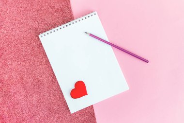 Boş sayfası olan bir defter, pembe arka planda kurşun kalem ve kırmızı kalp. Düz yatıyordu. Metin için yer. 