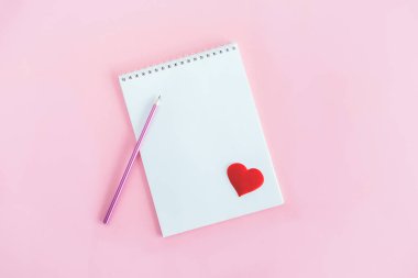 Boş sayfası olan bir defter, pembe arka planda kurşun kalem ve kırmızı kalp. Düz yatıyordu. Metin için yer. 