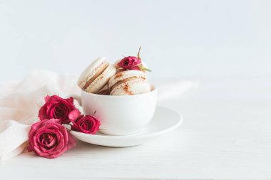 Beyaz fincanda pembe güller olan lezzetli Fransız kurabiyeleri. Beyaz ahşap arka plan. Metin için yer.