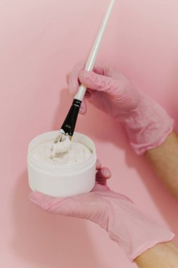 Beyaz kavanozda yüz maskesi ve kozmetik uzmanının ellerinde fırça. Pembe arka plan. Cilt bakımı güzellik ürünü. Kozmetoloji kavramı.