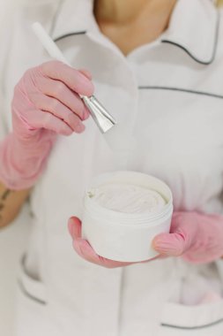Beyaz kavanozda yüz maskesi ve kozmetik uzmanının ellerinde fırça. Cilt bakımı güzellik ürünü. Kozmetoloji kavramı.