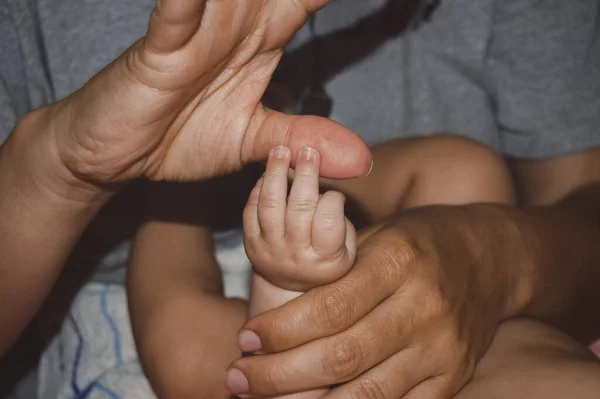 Yeni doğmuş bir bebeğin küçük elleriyle yetişkinlerin parmağını tutuşu. Şefkatli aile anları ebeveyn sevgisini, ilgisini ve korumasını gösterir. Yumuşak iç aydınlatma, samimi yaşam tarzı konsepti.