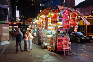 New York, New York, New York, ABD - 10 Aralık 2025: Turistler gece geç saatlerde Manhattan kaldırımının kenarındaki bir gıda standından fast food alıp yiyorlar. 