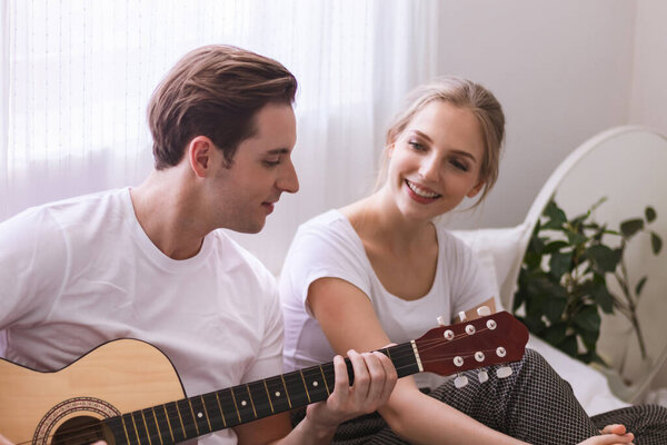 Lovely young caucasian couple or lover on bed together. Man play guitar to woman in dreamy romantic moment in bedroom. Photo of married relationship or bonding between boyfriend and girlfriend