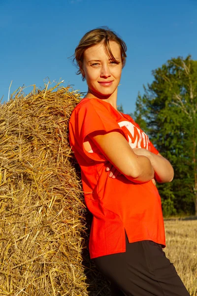 夕阳西下 一个穿着红色T恤的年轻漂亮姑娘在草堆和森林的背景下的画像 — 图库照片