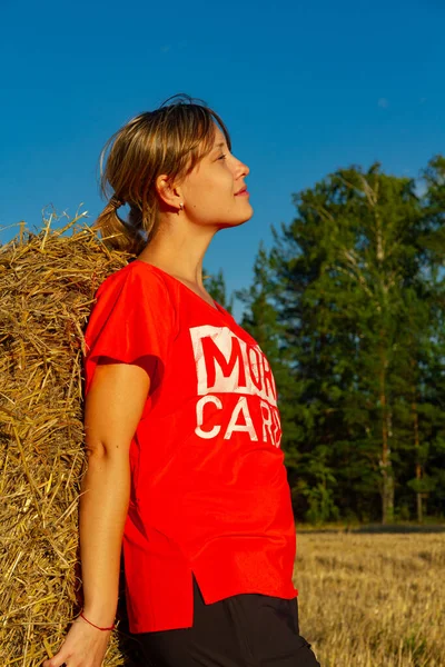 夕阳西下 一个穿着红色T恤的年轻漂亮姑娘在草堆和森林的背景下的画像 — 图库照片