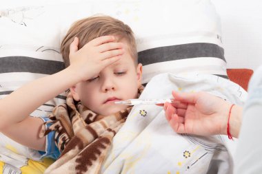 Küçük bir çocuk atkıya sarılmış bir çocuk evde hasta, yatakta yatıyor ve alnına tutunuyor, annesi ona bir termometre veriyor.
