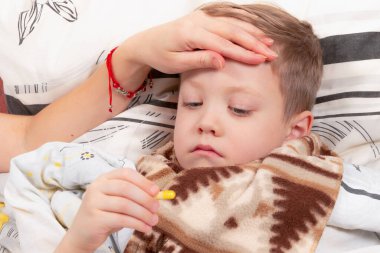 Eşarba sarılmış küçük çocuk evde hasta, yatakta uzanıyor ve ilaç içiyor.