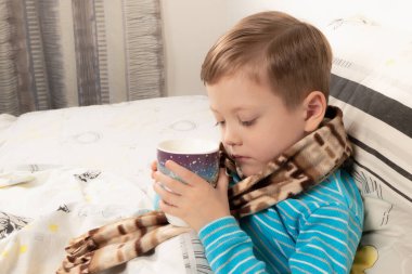 Mavi kazaklı hasta çocuk sıcak bir eşarba sarılıp ballı zencefilli çay içiyor.