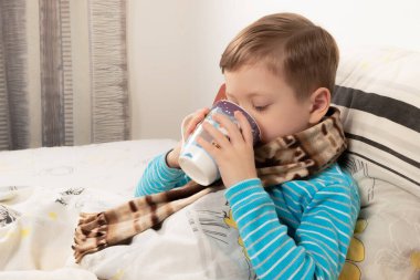 Mavi kazaklı hasta çocuk sıcak bir eşarba sarılıp ballı zencefilli çay içiyor.