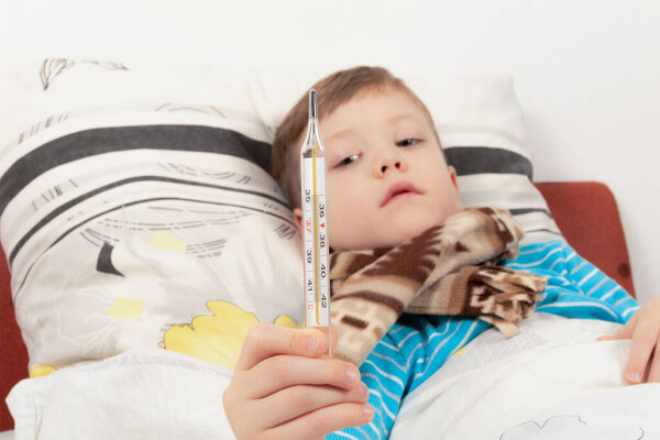 a sick child a boy in a blue jacket lies at home in bed and measures the temperature with a thermometer