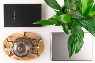 Ahşap tezgahta bitkisel çaydanlık, gri tablet, yeşil bitki ve defter beyaz ahşap masa üzerinde.
