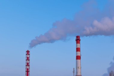 Bir fabrikanın kırmızı ve beyaz bacası. Güneşli bir kış gününde gri dumanlar mavi gökyüzüne karşı çıkar.