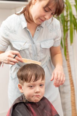 Açık mavi elbiseli, elinde tarak ve makasla güzel bir anne oğlunun saçını evde kesiyor. seçici odak