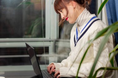 Sıcak kazaklı genç bir kız evdeki pencere kenarında dizüstü bilgisayarın başında oturuyor.