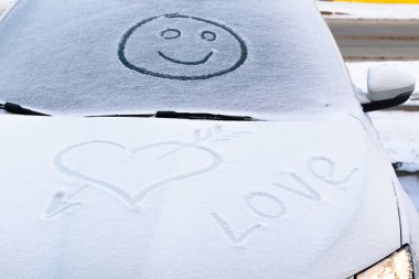 Karda gülümseyen bir yüz bir arabanın ön camında ve soğuk bir kış gününde kalp şeklinde.