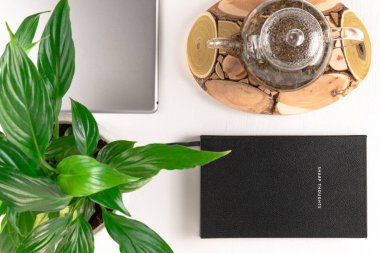 Ahşap tezgahta bitkisel çaydanlık, gri tablet, yeşil bitki ve defter beyaz ahşap masa üzerinde.