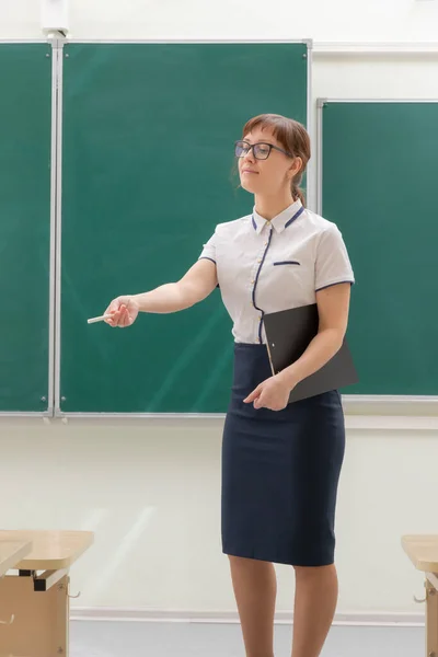 教室に緑の黒板の背景にフォルダを手にした白いブラウスと眼鏡の若い可愛い女性の学校の先生 — ストック写真