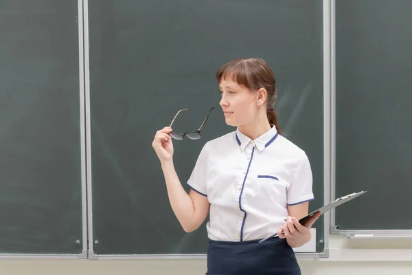 黒板の後ろの教室にフォルダと眼鏡を手に白いブラウスを着た学校の先生の若いきれいな女性 肖像画 — ストック写真
