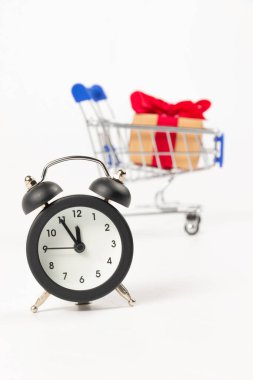 İçinde kırmızı bir fiyonk ve kurdele olan hediye kutusu. Tekerlekli, parlak bir alışveriş arabasında. Ve beyaz arka planda bir çalar saat. seçici odak
