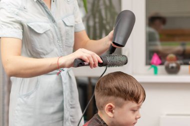 Elinde açık mavi elbiseli bir saç kurutma makinesi olan bir anne, karantinanın ikinci döneminde oğlunun saçını evde yapıyor. seçici odak