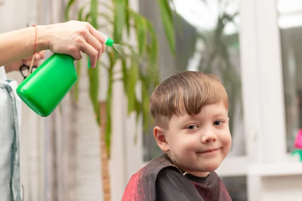 美容室の女性の手は 窓や緑の植物を背景に髪を切る前に 子供の頭の上に緑色のスプレーから水を噴霧します 選択的焦点 — ストック写真