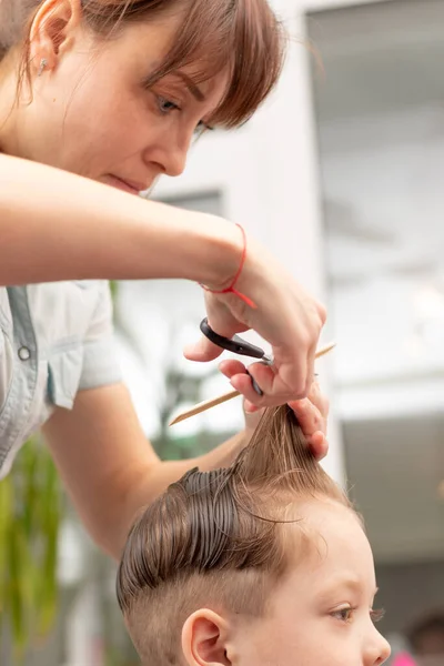 薄紫色のドレスに櫛とはさみを手にした若い母親が家で息子の髪を切ってる 選択的焦点 — ストック写真