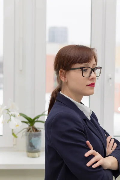 穿着蓝色夹克和眼镜的年轻而严格的商界女性正在工作场所的办公室橱窗前思考着 — 图库照片