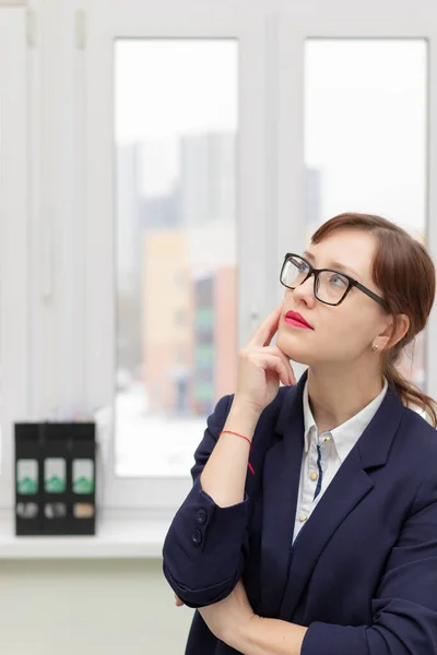 穿着蓝色夹克和眼镜的年轻而严格的商界女性正在工作场所的办公室橱窗前思考着 — 图库照片