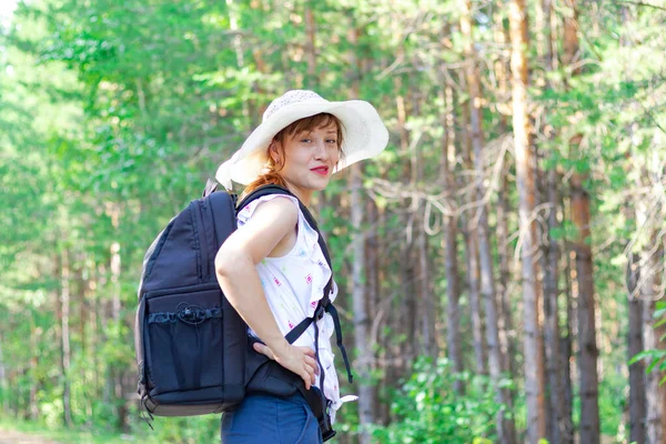 一个阳光灿烂的夏天 年轻的女游客背着背包 头戴草帽 站在绿树成荫的森林里 有选择的重点 — 图库照片
