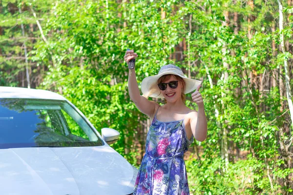 夏日的晴天 在绿树成荫的背景下 一位美丽而快乐的女人手握钥匙 站在一辆白色轿车前 有选择的重点 — 图库照片