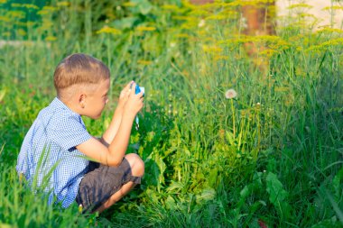 Mavi gömlekli hoş bir anaokulu çocuğu sıcak bir yaz gününde yeşil bitkilerin fotoğraflarını çekiyor. Seçici odaklanma. Portre