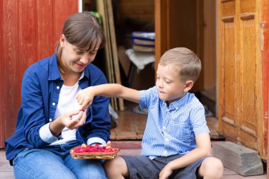 Altı yaşında bir çocuk annesiyle birlikte evin verandasındaki köyde taze kokulu ahududu yiyor. Seçici odaklanma. Portre