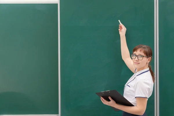 黒板の後ろの教室にフォルダとチョークを手に白いブラウスを着た学校の先生の若いきれいな女性 肖像画 — ストック写真