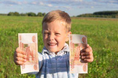 Tatmin olmuş bir çocuk, anaokulu öğrencisi bir çocuk, açık bir arka planda açık havada, güneşli bir yaz gününde yeşil çimenli bir alanda kağıt ruble tutuyor. Yakın plan. Portre