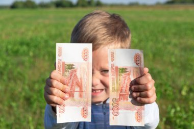 Tatmin olmuş bir çocuk, anaokulu öğrencisi bir çocuk, açık bir arka planda açık havada, güneşli bir yaz gününde yeşil çimenli bir alanda kağıt ruble tutuyor. Yakın plan. Portre