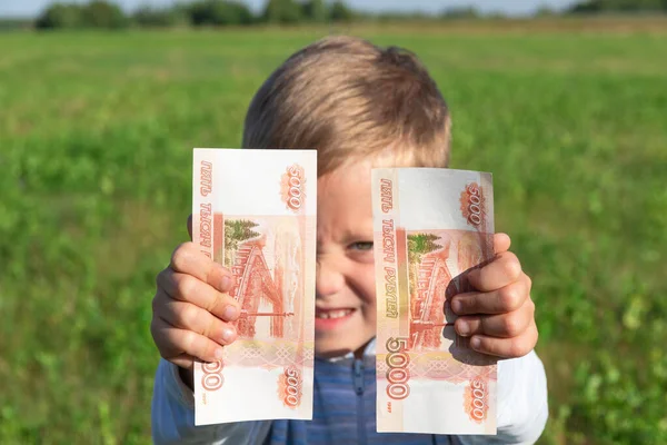 Tatmin olmuş bir çocuk, anaokulu öğrencisi bir çocuk, açık bir arka planda açık havada, güneşli bir yaz gününde yeşil çimenli bir alanda kağıt ruble tutuyor. Yakın plan. Portre