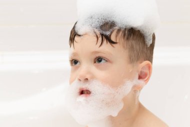 Kreş çocuğu açık beyaz bir küvette yıkanır ve köpükle oynar. yakın plan portre