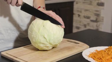 Kadın şefin elleri siyah mutfak masasındaki tahta kesme tahtasının üzerinde bıçakla beyaz lahana kesiyor. Yakındaki bir tabakta rendelenmiş havuç. Yakın plan.