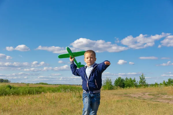 A six-year-old preschooler boy in a blue jacket launches a toy plane in a field against a blue sky with clouds on a summer day. The bright sun is shining. Scenery