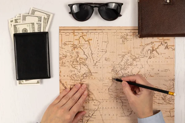 用铅笔的女性的手显示了世界地图上的位置 在一张白色的木制桌子上 放着一个装有钱 太阳镜和日记本的黑色皮夹 — 图库照片