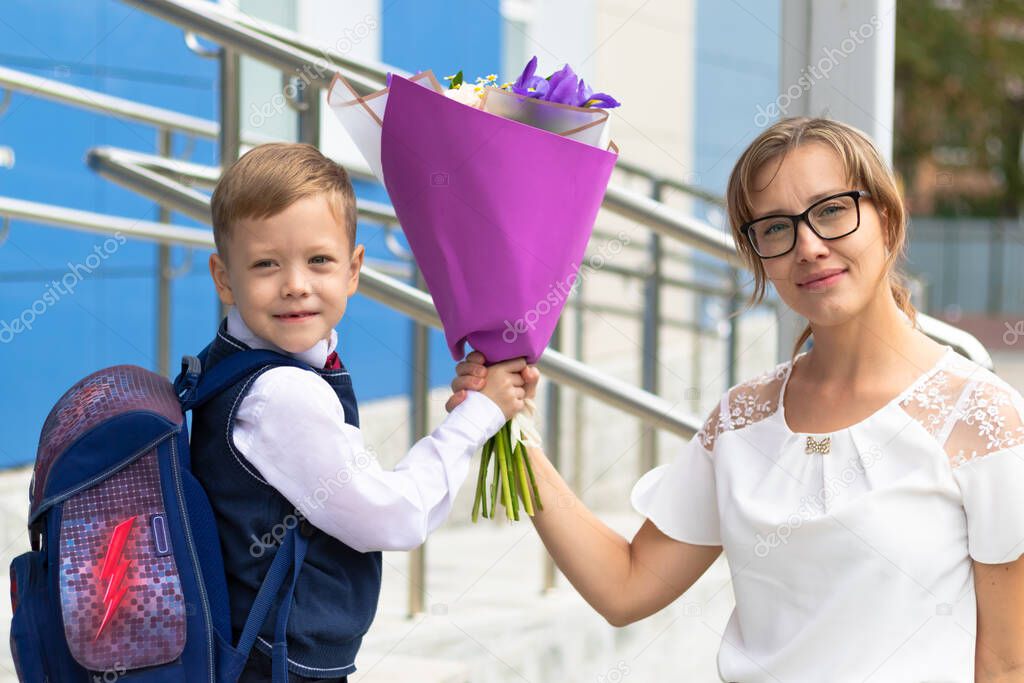 A cute first-grader boy in a school uniform with his mother and a beautiful bouquet of colorful flowers in the school yard. Celebration on September 1st. Knowledge day. Selective focus