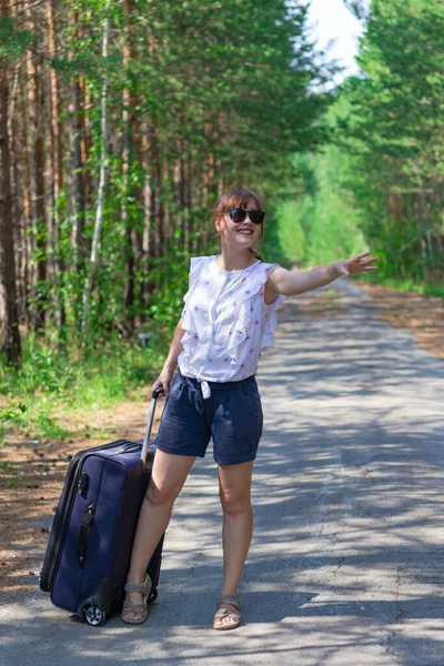 夏日阳光明媚的一天 在绿树成荫的背景下 一位漂亮的年轻女性游客提着手提箱走在林荫道上 有选择的重点 — 图库照片